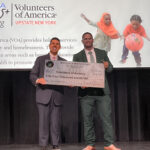 Dr. Junior Dillion and Senator Cooney pose holding a large presentation check on stage.